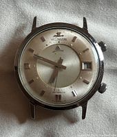 Straight-on front view showing silver sunburst dial, applied baton markers, date window at 3 o’clock, two crowns and acrylic crystal.