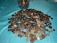 Image showing a large scattered pile of coins on a blue fabric surface, including a visible glass jar containing more coins.
