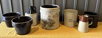 Photo of entire collection of 7 stoneware items including crocks, jugs, and pitchers on yellow cloth surface against grey wall.