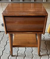 Front view of the nightstand showing drawer and tapered legs with metal caps, placed on patterned pavement.