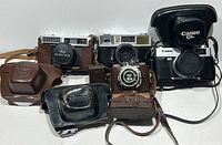 Front view of the four vintage cameras with leather and padded cases showing brand names on lens caps and cases.
