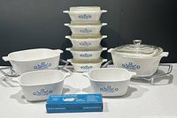 Front view of all CorningWare casserole dishes and accessories arranged on a table showing all sizes and lids, including metal cradles and lock-on handle.