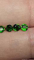Four round faceted intense electric green natural chrome diopside gemstones with excellent clarity shown on finger for scale.