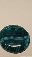 Close-up photo of a polished round striped agate cabochon showing multiple bands in green hues on a beige background.