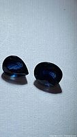 Two deep midnight blue sapphires side by side, one pear cut and one oval cut on a white background showing faceting and color.
