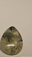 Polished teardrop-shaped natural mossy green prehnite gemstone with rutilated inclusions visible throughout the gem.