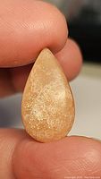 Close-up image of a pear-shaped peach moonstone cabochon held between fingers, showing its natural texture, color, and internal sparkle.
