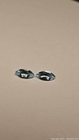 Two natural sky blue marquis cut topaz gemstones placed side by side on a beige background with visible facets reflecting light.