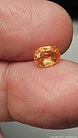 Close-up photo of a glowing mandarin sapphire held between thumb and forefinger showing oval cut facets and bright mandarin orange color with yellow undertones.