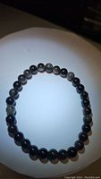 Photo of a black beaded stretchy bracelet with beads showing natural silver sheen reflections from obsidian stone, placed on a white circular background.
