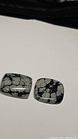 Pair of rectangular cushion-shaped Snowflake Obsidian cabochons showing typical snowflake patterns on black background, photographed on white surface.