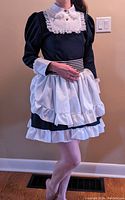 Front view of black Lolita maid dress worn by model, showing white bib with button, white apron, and ruffled skirt hem with striped sash tied at back.