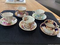 Top angled view of all five teacups with matching saucers on dark cloth, showing varied floral patterns and gold trim details.