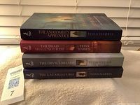 Four Tessa Harris mystery books stacked horizontally showing spine titles and colors: The Anatomist's Apprentice, The Dead Shall Not Rest, The Devil's Breath, and The Lazarus Curse.