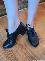 Front view of the black leather tap shoes being worn, showing lace-up design and shoe shape.