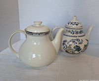 Photo of two ceramic teapots placed side by side on a white cloth background. Left teapot is cream colored with blue-gray pattern near lid, right teapot is white with blue, green, and yellow floral design and visible crazing.