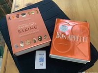 Two cookbooks lying on a dark cloth on a wooden table. 'Starting with Ingredients Baking' by Aliza Green is on the left, with detailed food imagery on the cover. 'Bon Appetit Cookbook' is on the right, with an orange glossy cover and a special offer sticker.
