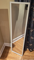 Tall thin mirror standing against a wall showing the full mirror and white plastic frame.