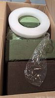 Photo showing a cardboard box with several green rectangular dry floral foam blocks and one white foam ring block.