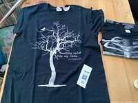 Black medium T-shirt with white graphic of a bare tree, a crow on a branch, and text reading 'Something wicked this way comes'. Shirt laying flat with tag visible.