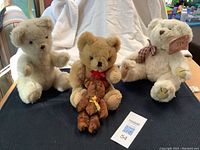 Photo showing three teddy bears standing and sitting, including one white, one light brown wearing red bow, and small dark brown teddy bear with yellow ribbon.