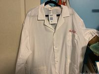 Photo showing front of white McGill lab coat with visible McGill red embroidery on chest pocket and button snapped up, hanging on hanger