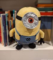 Front view of Stuart minion plush doll in yellow fabric with one large eye, silver goggle, blue overalls and black gloves/shoes.