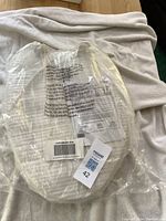 Elongated clay colored toilet seat still in its original clear plastic packaging with label and barcode visible.