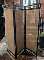 3-panel outdoor privacy screen showing black metal frame with decorative grid top and beige woven fabric privacy panels, standing outside on a paved area.