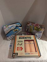 Lot displaying one unopened Throw Throw Burrito card game and two puzzle boxes, one opened with puzzle pieces, one unopened with collage artwork.