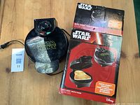 Closed Star Wars Darth Vader waffle maker with power cord and original branded box on a wooden table.