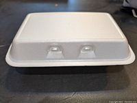 Close-up side view of a white styrofoam clamshell container closed, showing hinged lid and clousure tabs.