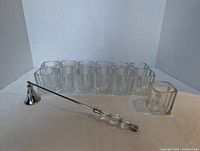 Thirteen clear glass votive holders arranged in rows with the chrome candle snuffer placed in front of them, on a white background.