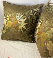 Photo showing two large square throw pillows with a dark green background and intricate floral and leaf patterns in yellow, white, orange, and green.