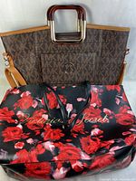 Two purses side by side. Front purse is Victoria's Secret with floral pattern, back purse is Michael Kors style monogram tote with wooden handles and tan strap.