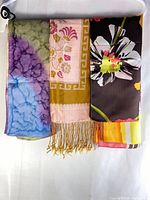 Three long silk scarves displayed folded side by side to show different patterns and colors including a batik-like pattern scarf, floral and Greek key bordered scarf, and a large flower patterned scarf.