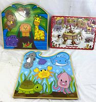 Three colorful wooden puzzles shown together on a white surface: a safari animal puzzle with monkey, lion, giraffe, hippo; an underwater animal puzzle with seahorse, whale, crab, squid, turtle; a Curious George circus scene puzzle with animals and people.