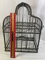 Full front view of dark silver ornamental bird cage with a red ruler vertically placed showing approximate height of about 16 inches.