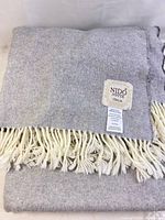 Folded gray wool throw blanket with visible Nido Notte label and cream fringed edges.