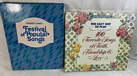 Two Reader's Digest music books side by side. One titled 'Festival of Popular Songs' in blue and purple cover; the other 'The Easy Way to Play 100 Favorite Songs of Faith, Friendship & Love' with a floral cover.