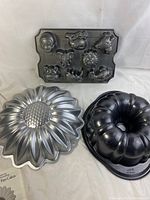 Three metal bakeware pans: a Nordic Ware zoo animal muffin pan with 8 animal-shaped molds, a Wilton sunflower cake pan with detailed petals, and a Wilton black bundt pan with fluted edges.