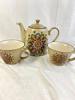 Off-white ceramic teapot and two matching mugs with colorful floral mandala pattern.