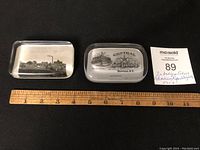 Two rectangular glass advertising paperweights positioned next to a ruler for size reference and a lot tag.