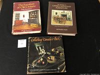 Three Canadian antique hardcover books sit on a black surface. Covers visible: a red book titled 'The Furniture of Old Ontario' by Philip Shackleton with image of room and furniture; a brown book titled 'The Heritage of Upper Canadian Furniture' by Howard Pain showing furniture; and a black book titled 'Collecting Canada's Past' showcasing a still life with fruit and bread.