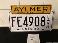 Front view of yellow Aylmer Ontario souvenir license plate topper attached to a white Ontario Farm license plate FE4908 with black lettering.
