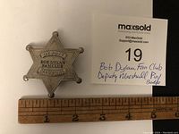 Front view of the star-shaped metal badge next to a ruler showing the badge is about 2 1/4 inches wide, with a note card describing the badge.