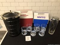 Lot displayed with all items: black plastic ice bucket with chrome lid on left, two Igloo coolers (red Little Playmate and blue Munchmate) in center back, six clear plastic Toronto skyline drinking glasses arranged in front of coolers, and a tall clear glass drink mixer on right.