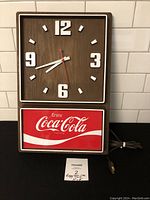 Full view of the Coca-Cola electric clock showing wood-tone face, white numerals, and illuminated Coca-Cola sign panel below.