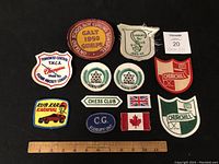 Top view of all 12 vintage crests arranged on a dark background with a ruler for scale. Various shapes, colors, and text are visible.