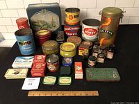 Wide assortment of 30 tins and containers arranged on countertop including coffee, honey, spice, and wax tins with some missing lids.